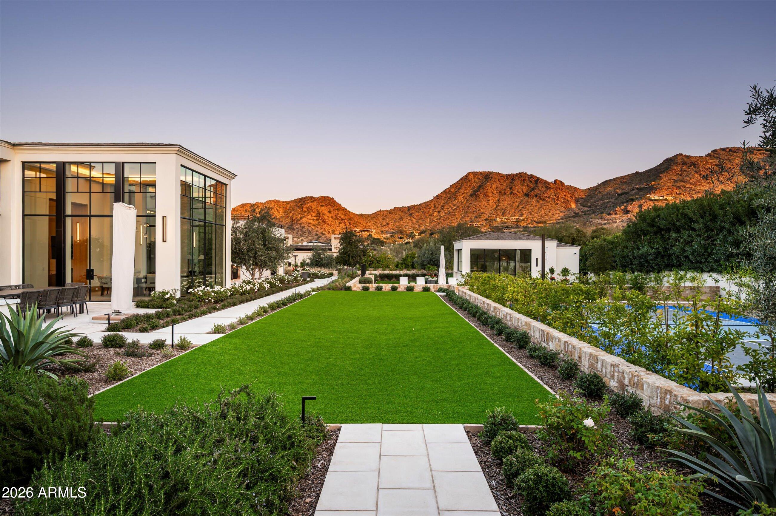 Set just north of Paradise Valley Country Club, Desert Park is a 2023 new build, move-in ready estate offering sweeping 180 degree views of Camelback, Mummy Mountain and Phoenix Mountain Preserve. Conceived by Candelaria Design, constructed with all steel framing by Arcadia Custom Builders, and finished with interiors by Katie Bowe Designs, the home reflects a rare level of craftsmanship and thoughtful collaboration. Lush grounds by Berghoff create a seamless connection between architecture and landscape. With iconic Camelback and Mummy Mtn. as your backdrop, the setting sun casts warm light across the landscape, creating a stunning contrast to the lush, green, flower-filled backyard. As you pull up to the home you will be impressed by the tall stately steel windows that open to a grand foyer. The main residence offers five bedroom en-suites, two powder baths, all designed with comfort and privacy in mind. Every room engages with the outdoors! At the heart of the home, is an atrium style dining room with soaring glass and stunning mountain views. A striking dual kitchen concept showcases Taj Mahal stone countertops with refined oak cabinetry, along with an additional gourmet prep area that includes a long center island, a full size Sub-zero freezer, a dishwasher, a prep sink, Wolf induction steam oven, convection oven, and Wolf double ovens. Adjacent to this room, is an oversized pantry and a commercial walk-in cooler.  A standout laundry room elevates everyday living with an island workspace, perfect as a wrapping station or craft counter, double stacking washers and dryers, built-in desk, and custom storage lockers.
The south-facing backyard is an entertainer's dream, featuring a diving pool and spa, expansive covered patios, manicured gardens, a second level view deck, a charming outdoor fireplace and Pickleball Court. A thoughtfully designed garden corridor offers a dedicated space ideal for flowers, herbs, or a seasonal kitchen garden, adding both beauty and functionality to the grounds.
A detached casita adds flexibility with its own kitchenette and living room, laundry, and spacious bedroom, perfect for guests, fitness, or a private studio. Dramatic 10ft telescoping steel doors open to the pool and entertaining area. The main gated entry of the home leads to two separate garages including a three bay garage with dog wash and a temperature controlled two bay garage. Both garages feature ceiling heights designed to accommodate car lifts, catering to the most discerning automotive enthusiast. This is truly a rare offering - an extraordinary, one of a kind estate that must be experienced to be believed! Furnishings can be negotiable.
