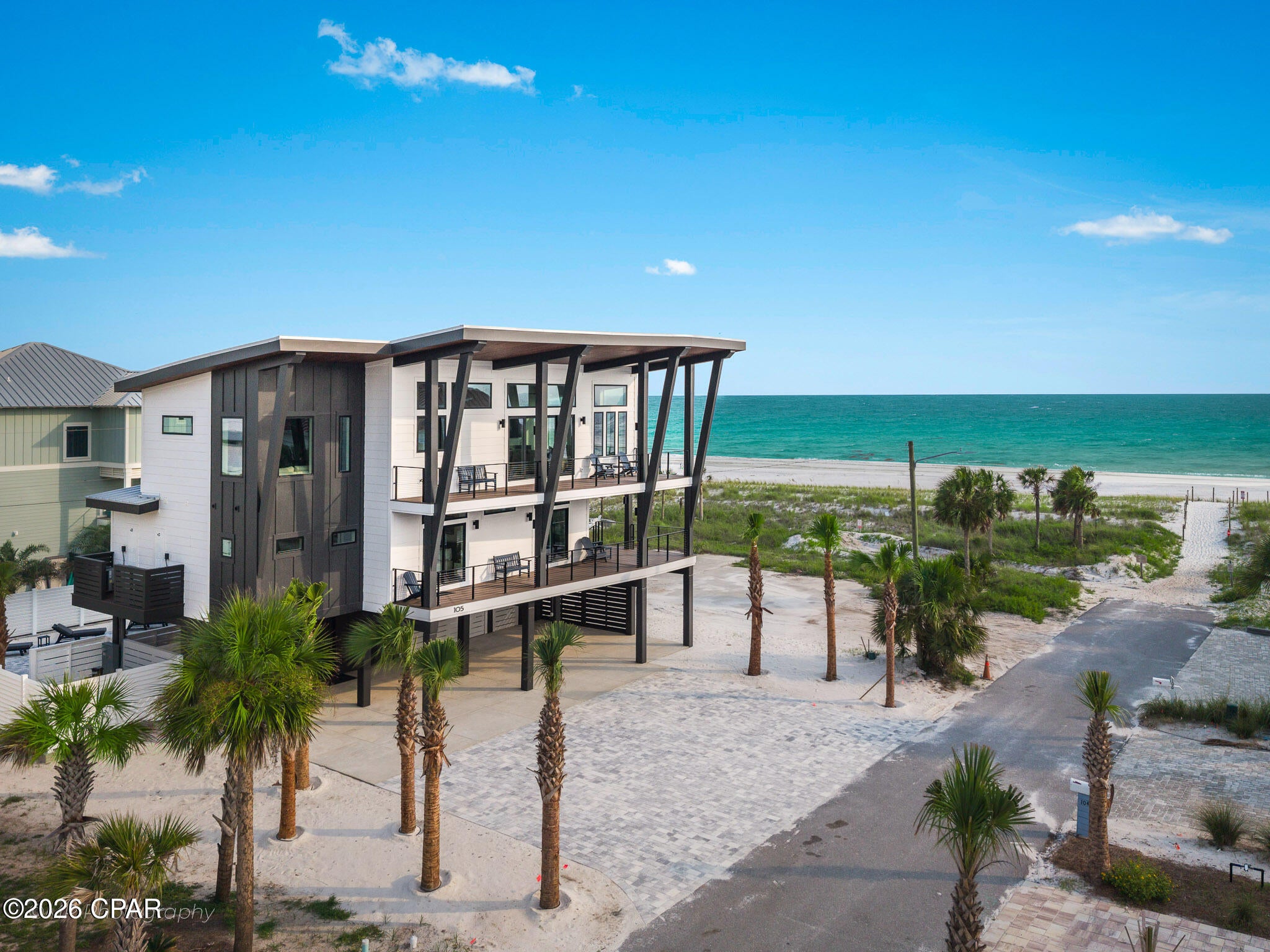 105 S 26th Street is a study in coastal architecture as much as it is a luxury residence, a brand new 5 bedroom, 5 bathroom home built around the logic of its site, second row from the Gulf in Mexico Beach. Long angled black steel columns support deep overhangs and a stained wood ceiling that carries across two full levels of balconies, each one looking straight out at the water.

An elevator runs to every floor. The view is never more than a button press away. Inside, the main living level opens around a waterfall quartz island, a custom range hood, and a Viking range, with the kitchen, dining, and living space sharing one wall of glass facing the Gulf. Five bedrooms and five bathrooms carry the same level of finish throughout the home, including a queen over queen bunk room built for family and guests alike. Four of the five bedrooms have private ensuite baths, each with its own custom tiled shower design. Upstairs, a dedicated game room with real arcade and pinball machines offers something few beach homes can claim.

Outside, a private pool and paver patio center on a stone fire pit, fenced in for privacy, with direct beach access just yards away at the end of the street. Just beyond the house, Mexico Beach unfolds at an easy pace, a short walk or golf cart ride to local restaurants, a coffee shop, and food trucks along the water, without the density or noise of a larger beach town.

Mornings begin with coffee on the balcony, watching the Gulf shift color with the light. Evenings settle around the fire pit, the sound of the waves close enough to hear. This is what buying on the Forgotten Coast is really about. Not just a house near the water, but a life built around it.