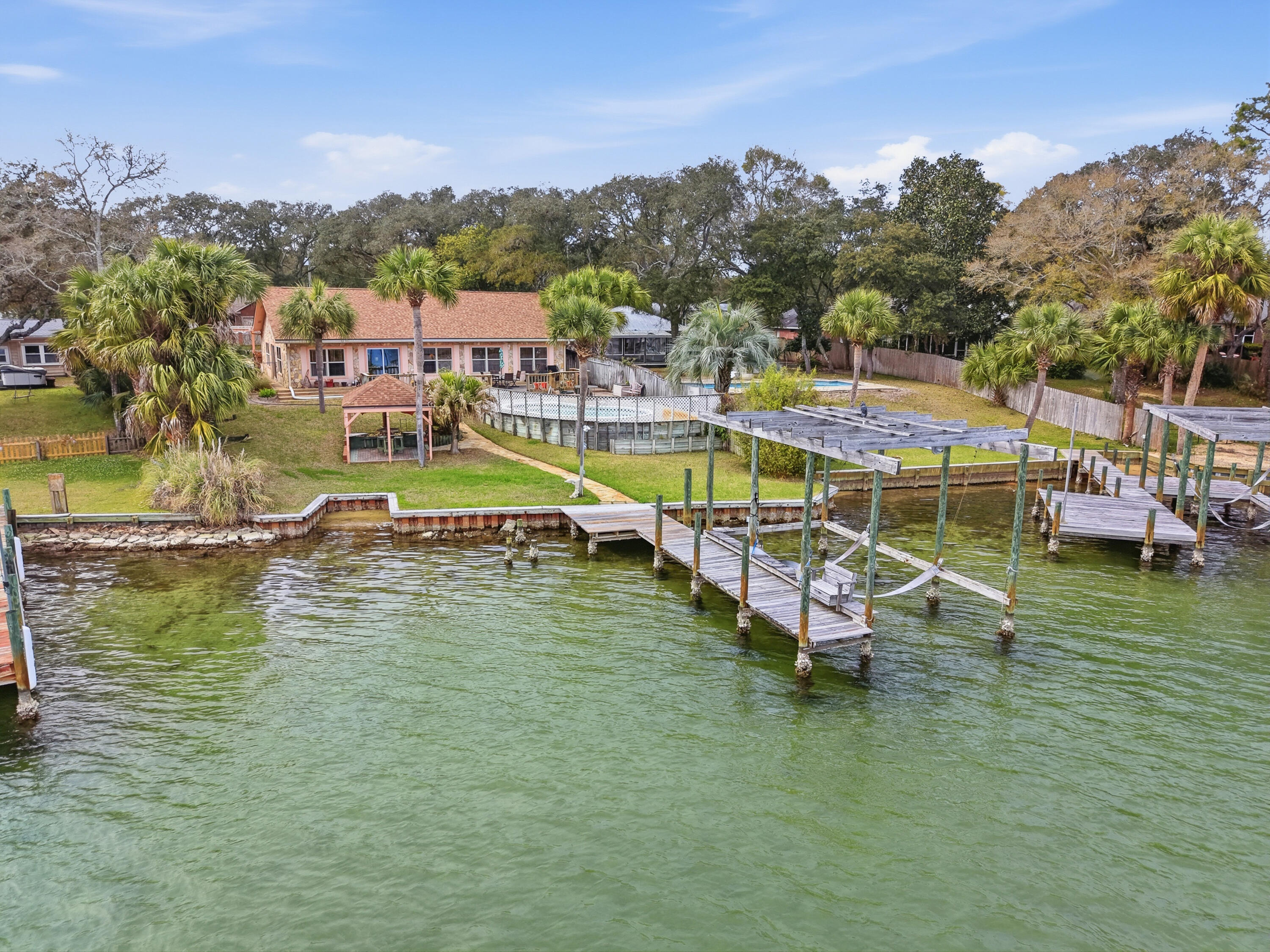 Positioned on ½ acre along the tranquil waters of Cinco Bayou, this exceptional waterfront estate at 144 Monahan Drive offers a rare blend of privacy, craftsmanship, and coastal lifestyle living. Designed for both comfort and entertainment, the property combines timeless finishes with distinctive architectural details throughout. Inside, expansive living areas showcase vaulted ceilings, cedar accent walls, built-in bookcases, and fireplace that create warmth and character. Travertine, porcelain tile, bamboo, Brazilian cherry, cork, and wood-style plank flooring reflect the custom quality found in every space. The chef's kitchen is appointed with built-in appliances and travertine flooring, flowing seamlessly into generous dining and gathering areas ideal for hosting. The waterfront primary The waterfront primary suite offers serene views and a spa-inspired bath featuring a soaking tub, bidet, oversized tiled shower, and custom walk-in closet. A private apartment lockout with skylights and bay window provides flexible accommodation for guests or multigenerational living. Outdoors, resort-style living awaits with a kidney-shaped in-ground pool, expansive paver deck, gazebo, and large wooden waterfront deck. Boating enthusiasts will appreciate the dock and lift (conveyed as-is), offering direct access to the bayou lifestyle. Additional highlights include a 12-12.5KW generator supporting major systems, whole-house water softener, detached 40x10 powered workshop, detached two-car garage, carport, and circular drive. This is more than a waterfront home it is a private coastal retreat in the heart of Fort Walton Beach, offering space, flexibility, and unforgettable sunsets over Cinco Bayou.