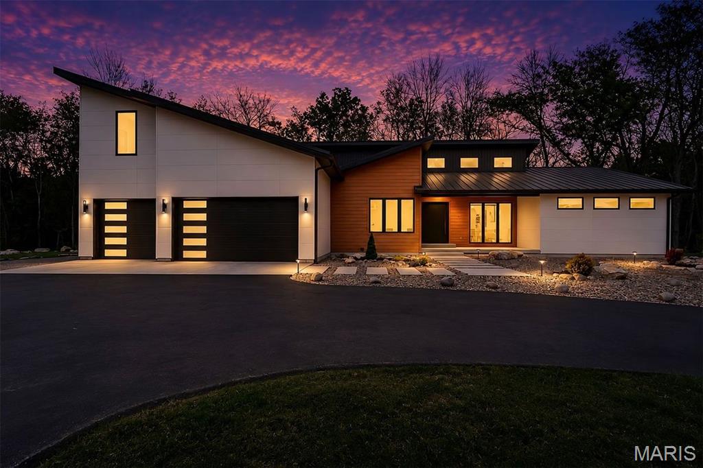 Set on more than four private, wooded acres, this brand-new Mid-Century Modern masterpiece redefines contemporary living with architectural precision and effortless sophistication. A long, dramatic drive leads to the striking facade, where clean lines, expansive glass, and impeccably landscaped grounds create a true sense of arrival. A stunning black metal pivot door opens to soaring ceilings and an open-concept plan bathed in natural light from high transom windows. The living room centers on a sleek linear fireplace, while the designer kitchen impresses with custom cabinetry, a sprawling island, and a built-in coffee bar for refined everyday living. The main-floor primary suite is a private sanctuary, featuring its own covered patio, dual walk-in closets, and a spa-worthy bath with a soaking tub encased in a glass-wrapped shower. A stylish laundry and drop zone complete the main level. Upstairs, a lofted office with a separate sitting area provides the perfect creative escape, while a breathtaking floating staircase with all-glass railings leads to the finished lower level, offering two bedrooms, a full bath, and expansive recreation space. Outdoors, mature trees and total privacy create an inspiring natural backdrop for future amenities or a peaceful retreat. A three-car garage and exceptional craftsmanship throughout complete this modern estate, where design, nature, and serenity exist in perfect balance.