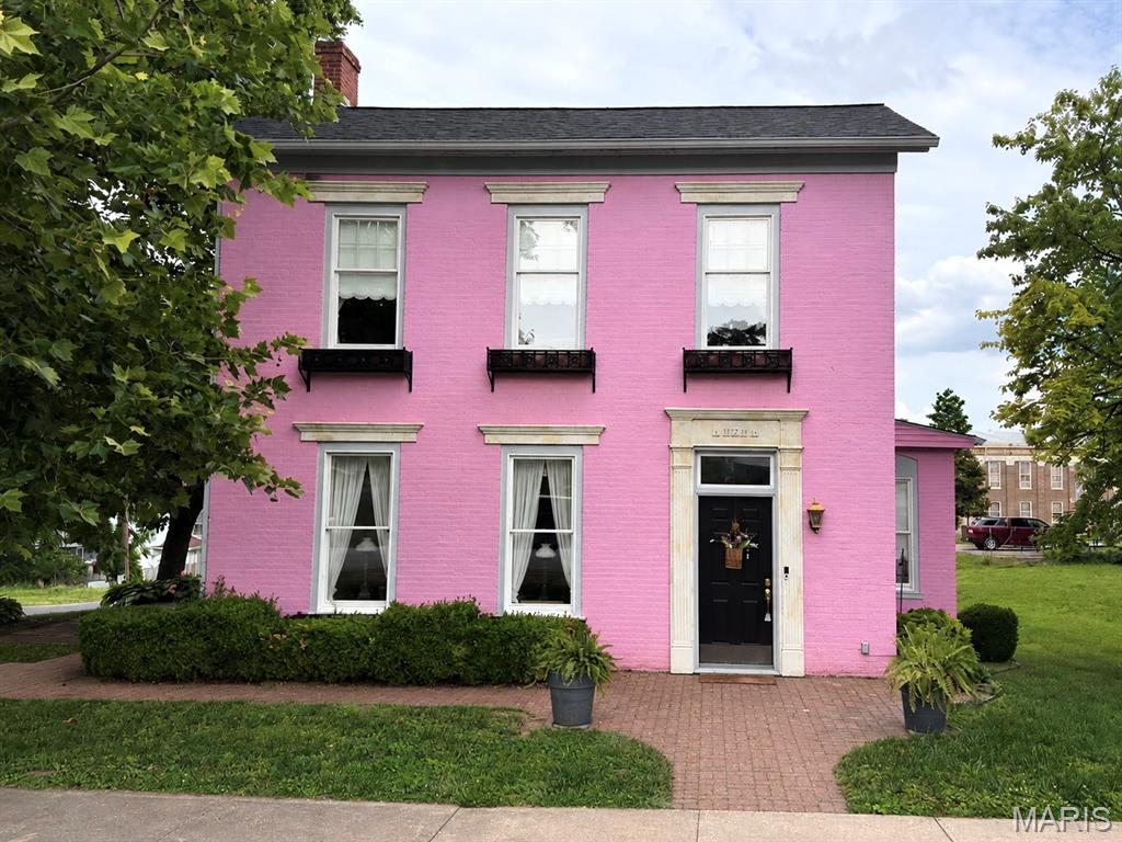 Imagine owning one of those rare homes that everyone recognizes—a home whose story has become woven into the fabric of the community.
When Mrs. Dottie dreamed of bringing a little of the charm and color of New Orleans to our quaint Mississippi River town, John lovingly embraced her vision. Together, they transformed this stately 1870 two-story brick home into the "Pink Lady.” 
Known for much more than its distinctive color, this home is is a reflection of John and Dottie - a couple devoted to their family and their community - deeply loved in return. They welcomed family, friends, neighbors, and visitors with warmth and genuine hospitality, pouring that same love into every thoughtful improvement they made to this remarkable home. Their passing leaves a void for many, but the care, joy, and pride they invested here remain evident throughout the property.
Inside, you'll find a home with timeless elegance complemented by carefully planned updates and meticulous attention to detail. 
Outdoors, the charm continues. Meandering brick walkways and inviting brick patios create welcoming spaces to gather, while thoughtfully designed landscaping frames the home in every season. You have handcrafted window boxes, beautifully upgraded trim and moulding to the custom doors, mailbox, and distinctive tile address plaques. A matching detached garage with garden workspace lean-to complements the home and large corner lot. 
John and Dottie left their mark not only on this remarkable home, but on the hearts of those who knew them. Their legacy lives on in every brick, every bloom, every carefully chosen detail, and every welcoming space they created. Now, The Pink Lady is ready to begin her next chapter with someone who will appreciate the love, craftsmanship, and history that make her truly one of a kind.