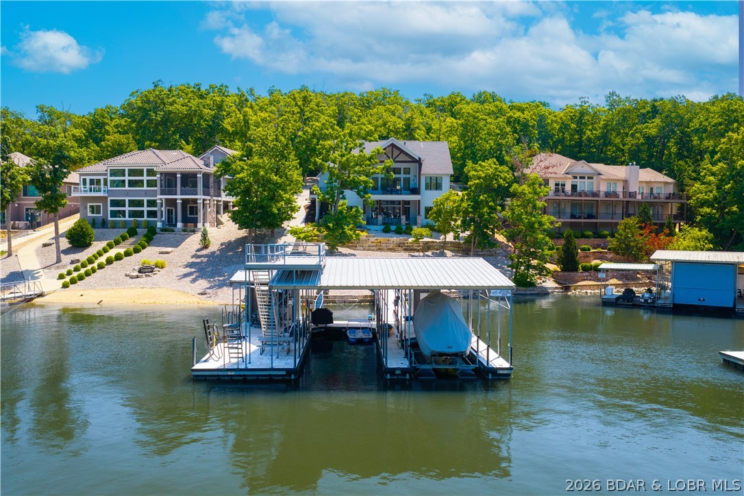 The ultimate Lake of the Ozarks retreat awaits in prestigious Porto Cima! Nestled in a deep-water cove, this custom Doug Ross home offers the rare combination of calm water for swimming, paddleboarding & family fun, plus year-round deep water and an incredible dock. With nearly 4,200 sq ft, 6 bdrs & 6 full baths, there's room for everyone to enjoy the lake lifestyle. The open main level features soaring ceilings, a floor-to-ceiling stone fireplace, a beautiful kitchen with quartz tops, the primary suite, guest bedroom & main-level living. Downstairs offers a spacious family room with wet bar plus 3 additional bedrooms, while the bunk room above the garage is perfect kids/guests. A golf cart path leads from the garage to the concrete dock featuring 2 boat slips, PWC spaces, a swim platform, bar & custom high dive that will quickly become a favorite. Multiple decks/patios overlook the cove, with the lower patio creating the perfect fire pit area. Modern finishes include black windows, cement-board siding, luxury vinyl plank flooring, remote blinds & abundant storage. If you've been searching for a luxury home where family & friends will gather year after year, this is the one.