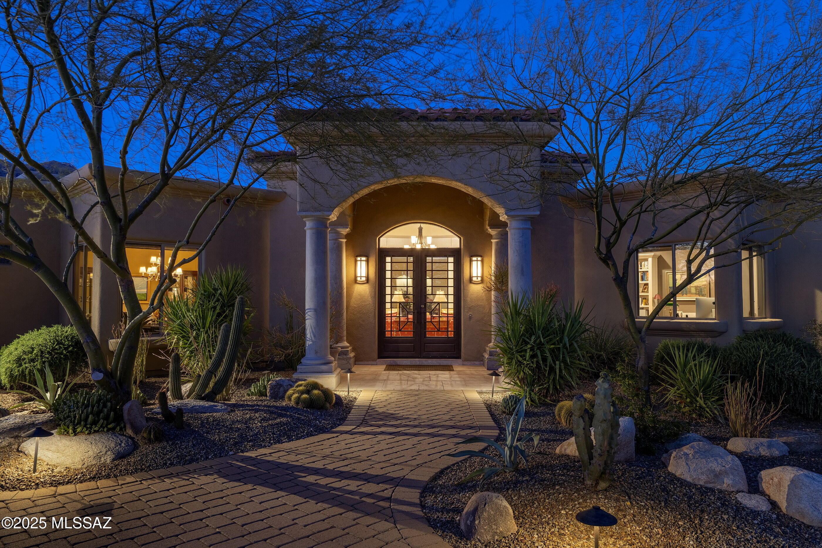 Nestled within the exclusive guard gated community of Pima Canyon, this thoughtfully designed residence embodies a timeless aesthetic, blending classic architectural details juxtaposed against the rugged beauty of the Sonoran Desert. The approach to the home unfolds as a carefully composed sequence, where the curated landscapes provide a textured backdrop to the residence's stucco clad façade and gracefully arched entryways. The interplay between structure and setting establishes a welcoming presence, creating a seamless transition between built form and natural environments. Stepping through the striking custom glass and iron entry, the interiors extend an invitation, spaces conceived not as mere enclosures but as experiences shaped by light, material, and craftsmanship. The living room, an expansive yet intimate volume, embraces the outdoors, framing the ever-changing drama of the Catalina Mountains through carefully positioned openings. Natural light is allowed to filter gently, shifting throughout the day and lending the space a dynamic quality.

The materiality of the interior is rich yet restrained, warm yet enduring. Italian Travertine flooring, handcrafted woodwork, textured walls, each element is imbued with a tactile honesty that speaks to craftsmanship and permanence. Wrought-iron and copper detailing punctuate the composition, offering moments of contrast and articulation, reinforcing the human touch within the greater whole.

The kitchen is designed with both beauty and function in mind, offering a refined yet approachable aesthetic that caters to both casual dining and formal entertaining. Custom Cherry wood cabinetry is complemented by hand-selected granite countertops, which provide a rich visual contrast. Design and utility converge in a space that is as visually stunning as it is effortlessly functional. A paneled Sub-Zero refrigerator, sheathed in warm wood to mirror the surrounding bespoke cabinetry, creates a seamless façade that speaks to the home's refined aesthetic. Just steps away, a spacious walk-in pantry offers discreet yet generous storage, while a central island, topped in honed granite, serves as an anchor for meal preparation.

Wrapping around the perimeter, an expansive secondary island with raised bar seating invites informal gatherings, offering a vantage point into the adjoining family room and reinforcing the kitchen's role as the heart of the home. At its culinary core, a professional-grade Viking cooktop is framed by a custom-designed hood, acting as a centerpiece enhanced by artisan-crafted tile work along the backsplash, an intricate, textural detail that elevates the room's cohesive, curated elegance. Adjacent to the kitchen, an intimate breakfast nook is bathed in morning light, offering a quiet retreat with views of the garden, while the formal dining room, with its statement lighting and classic wainscoting, provides a more structured setting for entertaining.

The residence's thoughtfully designed floor plan balances openness with privacy, creating distinct yet connected spaces. The owner's suite, privately positioned in the northeast wing, is a true retreat, offering a graciously proportioned bedroom with coffered ceilings, a fireplace for added warmth, and French doors that open onto a dedicated terrace. The adjoining bathroom features a freestanding soaking tub, a custom vanity with marble countertops, and a glass walk-in shower framed by hand-laid tile. Across the home, the secondary suites mirror the same attention to detail, with their own well-appointed bathroom and garden views that enhance the sense of retreat.

Beyond the interiors, a collection of outdoor living spaces extends the experience of the home, carefully positioned to maximize views while offering a variety of settings for relaxation and gathering. The rear veranda, shaded by deep overhangs, opens onto a series of tiered patios framing the pool, spa and outdoor barbecue. The arrangement is purposeful, positioning each element in relation to view, light, and movement. Native plantings and mature trees provide dappled shade, while meandering flagstone pathways connect the terraces, enhancing the sense of intimacy within the larger landscape.

Situated just minutes from La Encantada's premier shopping and dining, as well as La Paloma Country Club's renowned Jack Nicklaus-designed golf course, this residence offers a refined and comfortable lifestyle. Its seamless blend of traditional architectural elements, thoughtfully curated interiors, and connection to the surrounding desert landscape make it a home of enduring beauty and grace.