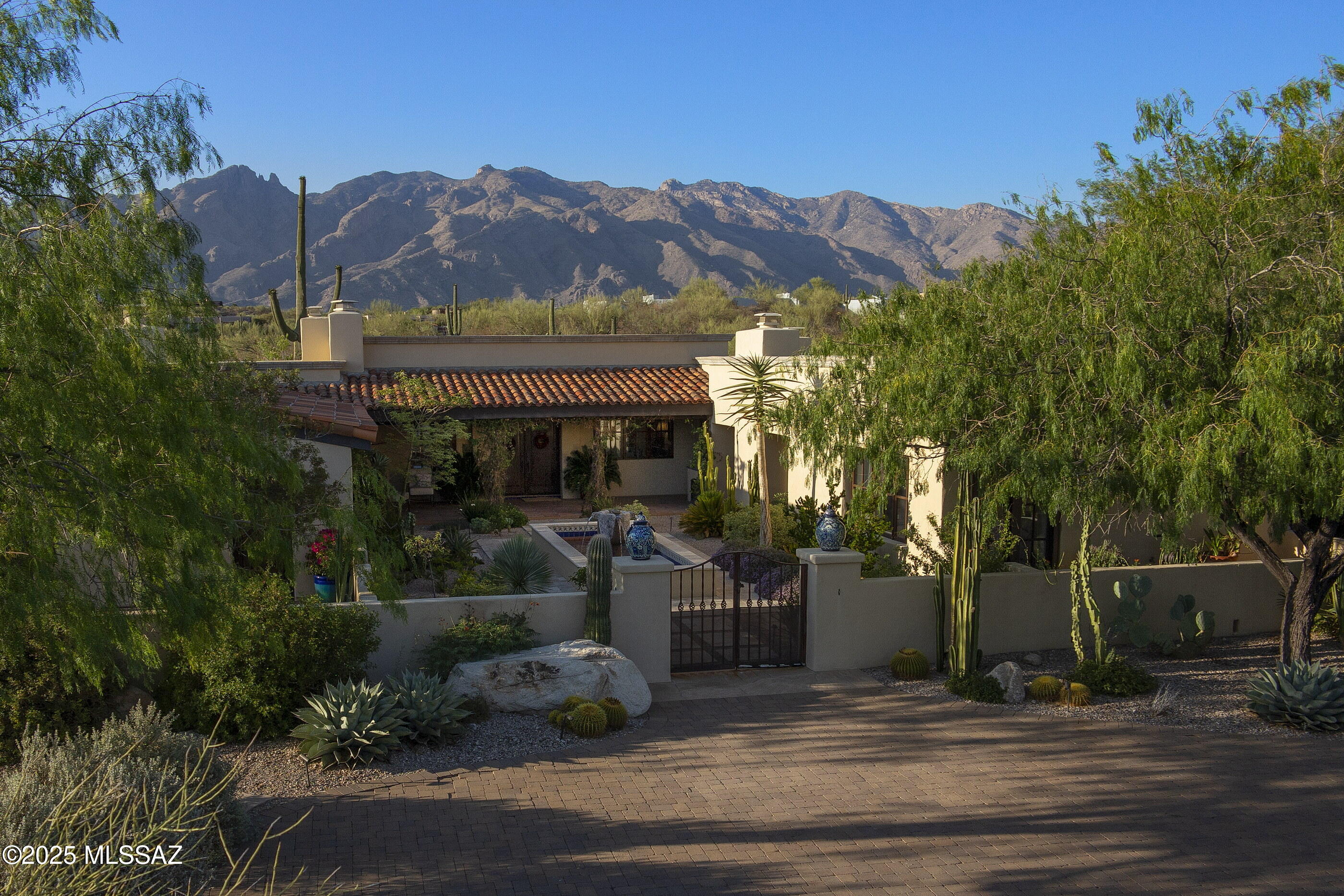 Gracefully positioned at the terminus of a quiet cul-de-sac within the distinguished, guard-gated community of Catalina Foothills 10, this private residence rests atop a 1.9-acre hilltop, commanding unobstructed vistas of the Catalina Mountains by day and a tapestry of city lights by night. Rooted in the regional architectural vernacular and inspired by the enduring soul of San Miguel de Allende, the home reflects a studied balance of old-world romance and regional expression. Approached by a gently winding paver drive, the home reveals itself through a formal motor court that opens onto an enchanting entry courtyard, centered by a tiered fountain and framed by an outdoor fireplace. The architecture unfolds in a classic central courtyard configuration, allowing nearly every principal room to engage with the outdoors through deep, shaded portals and view-framing arcades.

Spanning 3,349 square feet, the residence is a testament to thoughtful craftsmanship and timeless materials. Hand-hewn mesquite cabinetry, vibrant Talavera tilework, and exposed wood beams lend an air of authenticity, while every room is imbued with natural light and quiet grandeur. The interiors offer four bedrooms plus a dedicated office, arranged in a split plan to provide both privacy and harmony of flow.

At the heart of the home lies a warm and welcoming kitchen, anchored by a center island and a hearth-style fireplace. This gathering space opens effortlessly to a charming breakfast nook and a sun-kissed BBQ terrace, where meals and moments spill gracefully outdoors. The adjacent living and dining spaces are equally inviting, scaled for both quiet evenings and entertaining alike.

The primary suite occupies its own distinct wing, designed as a serene retreat that balances openness with privacy. A fireplace anchors the space, providing warmth and a subtle focal point without overpowering the calm atmosphere. Direct access to the spa creates a seamless connection between indoor comfort and outdoor relaxation. Adjacent, a custom library or study offers a quiet, well-lit workspace that supports both reflection and productivity. Generous in scale yet restrained in detail, the suite reflects a considered approach to living, where every element is intentional, materials are authentic, and a sense of calm prevails.

Situated on the opposite end of the residence, the secondary bedroom wing unfolds with an air of quiet grace, thoughtfully arranged around a flexible central room that invites relaxation, study, or intimate gatherings. Each of the two bedrooms opens directly onto its own private courtyard, a secluded outdoor sanctuary defined by stucco walls and lush landscaping, where morning light filters gently through leafy canopies and cool breezes stir the air. These courtyards, extending the living space beyond walls, embody the seamless indoor-outdoor living so integral to the Spanish Colonial tradition. The bedrooms themselves are generously proportioned, with tall windows and doors that frame serene views and invite the natural world in. This wing offers a harmonious balance of privacy and connection, creating a peaceful retreat for family or guests alike.

A rare offering of architectural integrity and enduring charm, this residence is more than a home, it is a legacy property, artfully composed and ideally situated within one of Tucson's most esteemed communities.