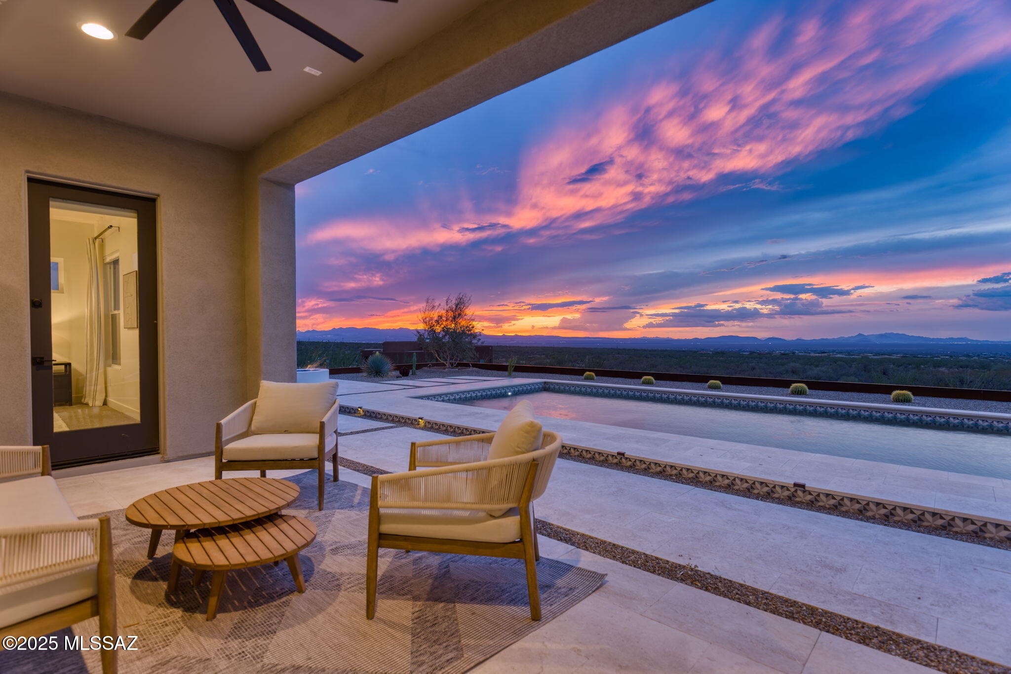Perched on 4.1 pristine acres in the ocotillo-strewn foothills of the Santa Rita Mountains at the edge of the Coronado National Forest, this 2023 soft-contemporary retreat captures sweeping, unobstructed views of the surrounding mountains and valley below. From its elevated vantage point, experience Tucson's iconic sunsets, endless star-filled skies, and sparkling city lights. With clean architectural lines and modern finishes, this private sanctuary is designed as a place to reflect, rejuvenate, and recharge.
A long driveway leads to a limestone walkway and covered entry, where a custom contemporary glass front door sets the tone. Inside, a wall of windows frames the uninterrupted views while filling the interiors with natural light. Continued...