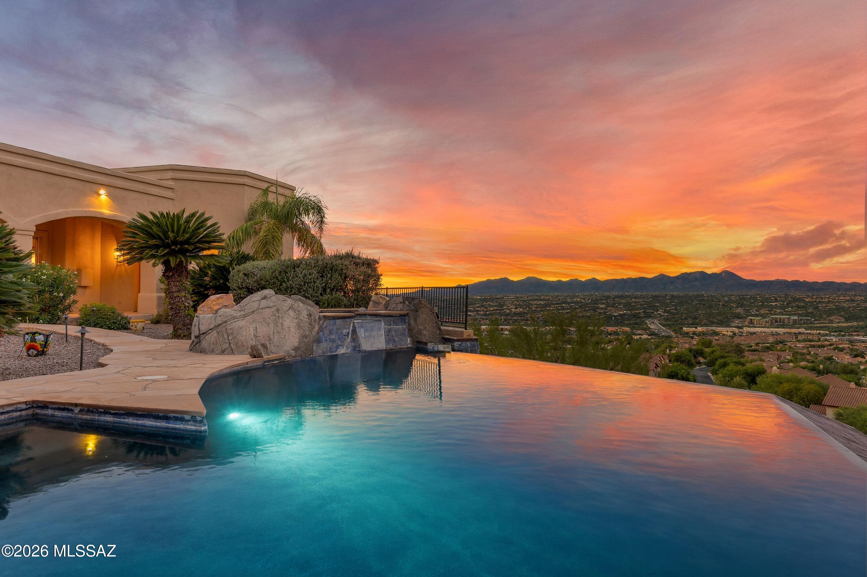 Perched on nearly two private acres in the prestigious gated community of La Reserve, this exceptional hilltop estate offers more than 5,000 square feet of refined desert living, with breathtaking views from nearly every window. Designed to embrace its dramatic setting, the five-bedroom, three-and-a-half-bath residence captures sweeping Pusch Ridge and mountain vistas by day and a dazzling panorama of city lights after sunset. Outdoor living is extraordinary. An expansive balcony provides a spectacular vantage point for enjoying the city below, while an immense covered rear patio and separate gazebo frame magnificent views of Pusch Ridge and the surrounding mountains. The negative-edge PebbleTec pool and spa create a true resort experience, seemingly suspended above the city (continued) lights and Sonoran Desert landscape. Inside, an ideal split-bedroom floor plan balances elegant entertaining with exceptional privacy. Gracious formal living and dining rooms set the tone, while the heart of the home is an expansive chef's kitchen appointed with luxury stainless-steel appliances, a substantial prep and dining island, a second island ideal for serving, and a large walk-in pantry. An oversized dining nook, surrounded by incredible views, comfortably accommodates everyday dining for up to eight. The kitchen opens seamlessly to the generous family room, where a wall of glass doors draws the desert landscape indoors. An adjacent full cocktail and wine bar makes this an effortless setting for gatherings both intimate and grand. For the ultimate entertaining experience, descend to your own private "speakeasy": an extraordinary 1,600-bottle wine cellar and tasting room tucked discreetly behind a privacy-glass door. A beautifully appointed powder room is conveniently located nearby, creating a destination within the home designed for memorable evenings and serious wine enthusiasts alike. The generous primary suite is a private retreat with direct patio access for enjoying sunsets, city lights, and easy access to the pool and spa. An elegant fireplace adds warmth and ambiance, while two sizable closets and an expansive primary bath feature separate vanities, a soaking tub, and a separate shower. The bath also opens directly to the outdoor living and pool area for a true resort-style connection. A fifth bedroom, currently used as an office, is conveniently positioned near the primary suite. On the opposite side of the residence, family and guests enjoy exceptional privacy with a separate ensuite bedroom and bath, plus two additional generously sized bedrooms and another full bath. An oversized, extra-height three-car garage easily accommodates vehicles while providing abundant additional storage. Private and serene yet remarkably convenient, this La Reserve estate offers an enviable combination of commanding hilltop views, expansive living spaces, and exceptional indoor-outdoor entertaining all just minutes from shopping, dining, hiking, and the amenities of Oro Valley and Northwest Tucson. A distinctive desert retreat where Pusch Ridge provides the daytime backdrop and shimmering city lights take center stage after dark.