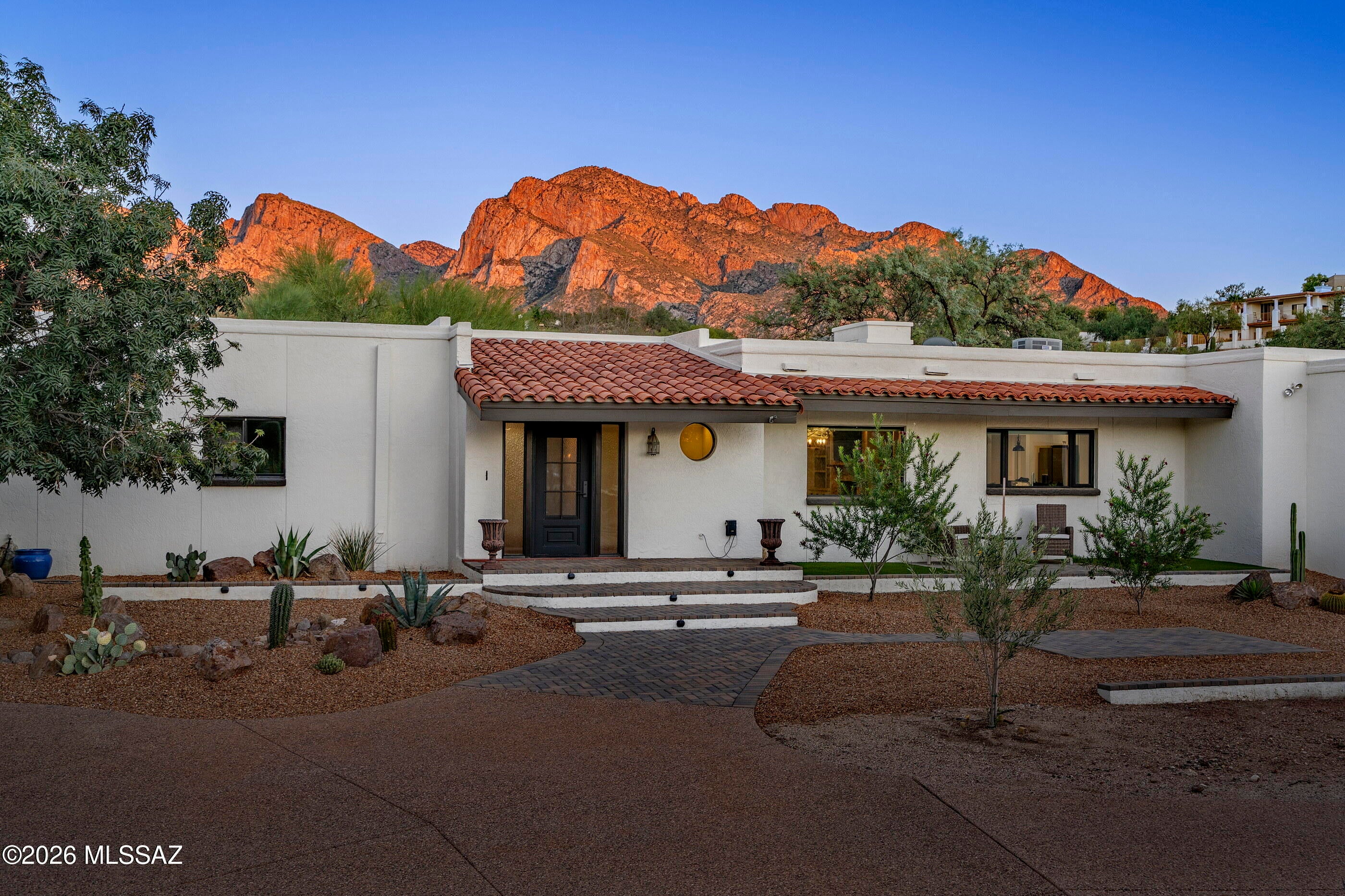 Located on one of the largest lots in the historic Oro Valley Country Club, this four bedroom home boasts a gorgeous interior and a six-car garage! Striking the perfect balance of modernity with vintage charm, this home sits on a 1.22 acre lot and has buildable space for a guesthouse.
A winding, circular driveway leads to the solidly built, masonry stucco home that boasts a freshly redone elevated ''porch'' with faux grass. A stylish, new steel door at the entry has an openable window with screen for added ventilation. Inside, the wide entry has space for a little sitting nook or ''drop zone''. A convenient half-bath off the entry has a dramatic tile feature wall in muted creams and taupes. Freshly painted in buttery cream and complimented by warm wood floors running throughout the home, interior of this home feels incredibly inviting. The main living space is open to the kitchen/dining and features a stacked stone fireplace with a quartz slab hearth, a beamed ceiling, a slider to the patio and built-in storage with display shelves. A walk-in closet off the living space has so many possibilities--extra storage or you could open it and create a study spot, dry bar or mini library. The generous dining area with room for a large table is highlighted by an 8-light farmhouse chandelier and a large picture window provides dinner guests with a pretty vista. The kitchen is a dream with all stainless steel appliances including a six- burner gas range with dual oven capability, pot filler and a steel hood. A large island with robin's egg light blue cabinetry anchors the space and has industrial farmhouse pendant task lights that make meal prep a breeze. The rest of the cabinets are cream color, and all are topped with veined quartz counters finished with ogee edges. The chic elongated hex backsplash is accented by floating wood shelves, while a vintage pantry cabinet adds even more storage and charm to the space. The cabinetry and counters in the kitchen are continued into the adjacent laundry room, which also has another pantry closet and extra fridge. Tucked around the corner from this area is a guest bedroom with easy access to the outdoors. This private bedroom with closet would make an excellent home office, gym or studio space. A large flex room leads to the primary and could be used as a second living space--or the new owner could add barn doors and enjoy it as part of the primary suite (office, sitting room, exercise room, create extra closet space, etc.). Both this room and the primary bedroom have sliding glass doors leading to brick-paved patios in the lush backyard. Set apart from the other bedrooms, the primary suite feels like a quiet sanctuary and has a stylish full bath that includes a wraparound shower, soaking vessel tub, dual sinks and a "smart toilet". The new owners will easily stay organized with the "California Closet" built-ins in the walk-in closet, which also features a hidden floor safe. There are two additional generous guest bedrooms serviced by a hall bath. One of these  guest bedrooms has a square panel feature wall and both bedrooms have plenty of closet space. The dramatic guest bath feels like a luxury spa with its fully tiled walk-in shower and feature wall in rich cocoa and sand tones. The shower's full-length linear niche, the floating wall toilet and heated towel bar all add style and functionality, while the creamy, stone-topped vanity with undermount sink and wood framed mirror tie the design together beautifully. Whether your goal is to entertain or relax, you will fall in love with the different outdoor spaces around the property. The large backyard is fully walled and offers brick paved patios, faux grass and mature trees. A fenced pool and spa area ramps up the fun factor with a vintage diving board and you can practice your putts on the green next to the raised paver stone under shady trees. Another paver patio toward the side of the home has banco seating and is accented by a large swath of green faux grass. Finally, an attached split six car garage/workshop with a storage room, cedar closet and EV charger round out this exceptional property. Valoro Estates is one of the only neighborhoods in the Oro Valley Country Club that does not have an HOA. (*This is a human written description from a human who has been inside the home. :))
