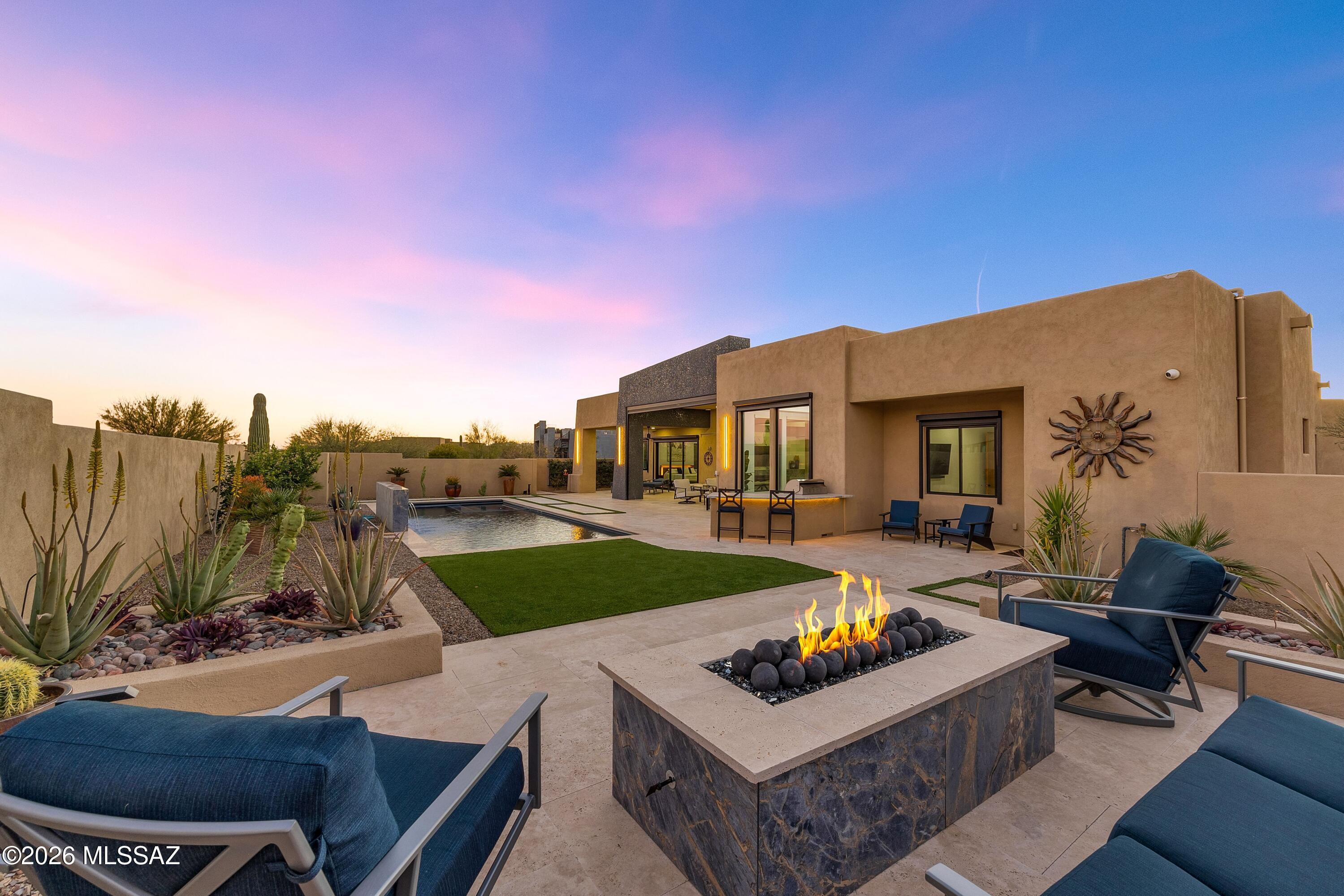Perfection awaits at 11931 N Mesquite Sunset Place; a stunning property recently featured in Tucson Lifestyle Magazine. The vision of a custom home builder, interior design team and landscape architect have come together creating a brilliant experience indoors and out. The home is conveniently located in a small, gated enclave in Oro Valley. Built in 2023 and enhanced thereafter, you will find every bell and whistle imaginable. Leading to a private entry, the paver drive has been finished with low voltage lighting and thoughtfully placed desert plantings. Residents enjoy an angled oversized 3 bay garage for vehicles and storage, while guests are greeted by a calming fountain & stunning glass pivoting door. Situated with eastern and western exposure, the home is positioned to take in both sunrise and sunset, with views of multiple mountain ranges.

Inside the 3,590 sq ft residence, high ceilings and light filled spaces are finished with high end windows, solid cherry doors, luxe quartz counters and custom cabinetry. A dramatic feature wall of gorgeous stone anchors the space and acts as a platform from which color is pulled into other spaces. Skylights are dotted throughout adding daytime illumination, while integrated lighting above soffits adds ambience at evening. Functionally designed with a split floor plan, the property offers 4 bedrooms, 3 ½ baths, plus a spacious den. Electronic blinds have been strategically placed both indoors and out to allow for light control, while maintaining views. Each bedroom has an ensuite or jack & jill bath, as well as a walk-in closet. Throughout, the condition is impeccable.

The indoors and outdoors unite with cascading sliding glass doors in both the great room and primary suite. With the help of multiple experts in outdoor design, the eastern facing backyard has been flawlessly finished. This space is truly an extension of the home. Flowing seamlessly, a spacious, covered patio with large format travertine extends to the heated saltwater pool and spa. The linear and modern pool was designed to include a water wall and an electric retractable cover. An outdoor kitchen features a built-in grill, fridge, granite countertop and bar seating. Positioned for sunset views, an elevated seating area has been finished with a gas firepit and built in planters.

This is not just a beautiful home -- it's an intelligent one. The owners have incorporated a full-home security system, smart controls and hard-wired cameras. Two EV chargers are tucked neatly in the garage, which features its own climate-control system via a Samsung/Lennox mini-split and epoxy flooring. Ample solar panels on the roof for low energy bills (pre-paid solar lease), a whole-house water purification system and reverse osmosis drinking water underscore an emphasis on sustainability and attention to wellness.

This location in Oro Valley is ultra convenient. Immediately outside the gate, runners and cyclists enjoy access to The Loop multi-use paved trail system. Within 2 miles, there is a large grocery store, dining, shopping and the Oro Valley library. Within 10 minutes, you will find Oro Valley Hospital, Oro Valley Marketplace and Catalina State Park. An incredible lifestyle awaits.