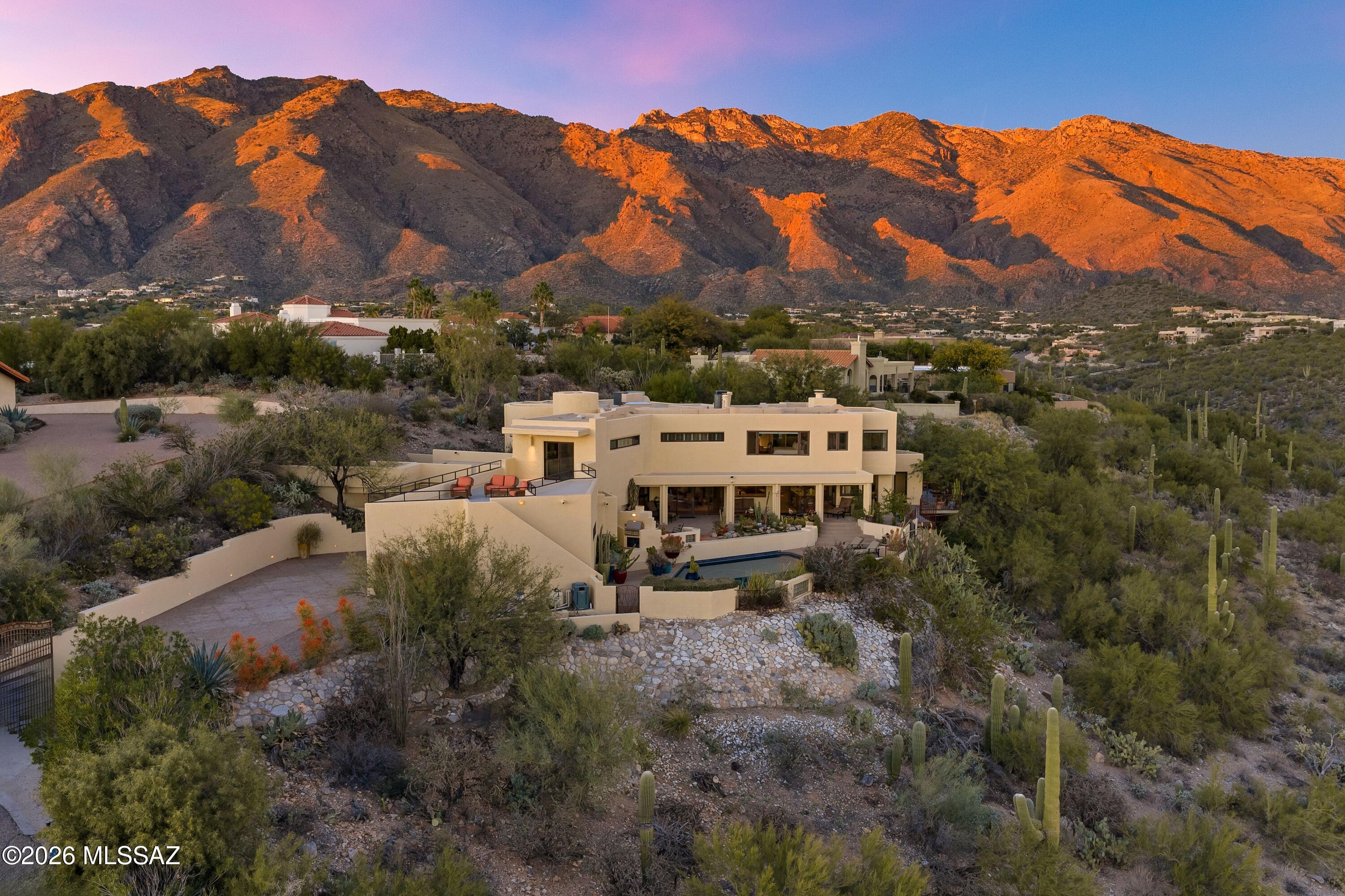Perched atop a ridgetop in Cimarron Foothills Estates, this exceptional Catalina Foothills residence commands some of the most spectacular views in Tucson, showcasing sweeping city lights and multiple surrounding mountain ranges. A cascading architectural fountain welcomes guests at the front entry, setting the tone for the refined experience that unfolds throughout the home. Travertine floors flow throughout light-filled living spaces, where thoughtful design balances comfort and sophistication. The expansive upstairs family room opens directly to a panoramic view deck, seamlessly extending the living space outdoors and creating an ideal setting for both intimate evenings and large-scale entertaining. Downstairs, a cozy living area is anchored by a rock-faced fireplace, (continued) offering a warm and inviting retreat perfect for relaxed gatherings or quiet moments. The dining room has a dramatic 2-story ceiling height and overlooks the walls of sliding glass doors looking onto the city view. The recently renovated kitchen is a true centerpiece, designed for the serious cook with high-end appliance appointments, dual islands, and an adjacent bar area. A third oven and a second refrigerator located in the three-car garage support effortless large-scale entertaining. Multiple patios provide a variety of settings for gatherings, complemented by a large PebbleTec pool overlooking the city lights, a long covered patio with adjacent outdoor fireplace seating, and a private spa nestled within serene northeast-facing gardens. The terraced north yard offers multiple seating areas framed by outstanding mountain views. Two primary suites provide exceptional comfort and privacy, including a beautifully upgraded main primary bath with an expansive walk-in shower and a freestanding tub, and an elegant vanity with glass storage cabinets. A downstairs guest suite with a dry sauna in the adjacent bath, along with two additional bedrooms and another bath, is ideal for family, guests, or office use, completing this remarkable foothills home, and an elevated entertainer's retreat defined by architectural detail, refined luxury, and extraordinary views.