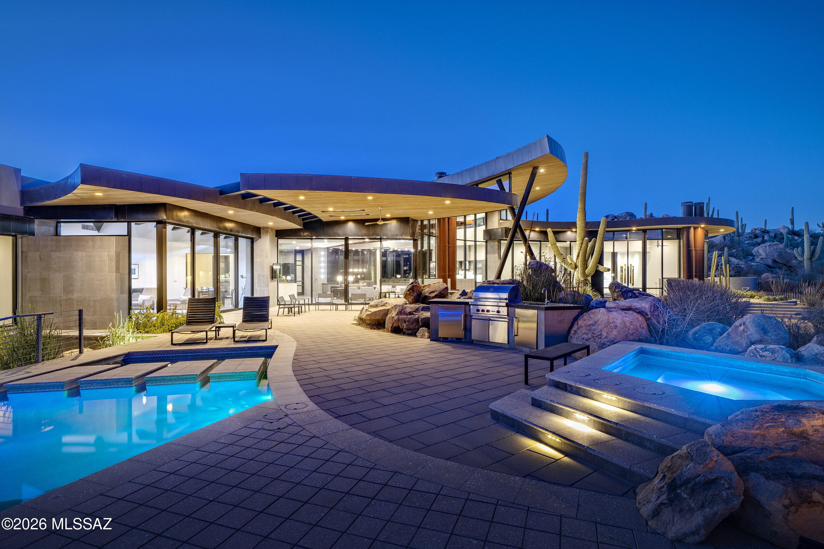 This Desert Modern estate, designed by renowned architect Marc Soloway with interiors by Lori Carroll, is gracefully nestled into the rugged boulder walls of Stone Canyon, standing as a poetic expression of architecture in harmony with its environment. The Southeast facing residence maximizes iconic views of Pusch Ridge and the Catalina Mountains while embracing the organic contours of this 1.9 acre parcel. The house reads as an extension of the very geology that embraces it: finely crafted Italian tile laid in striated patterns that echo natural stone formations, and custom metal surfaces patinate in desert sun, forming a nuanced palette of ash, sienna, and iron that reflects the surrounding boulders. Extensive overhangs, articulated through segmented and cantilevered planes, not only deliver a dynamic sculptural presence but also mitigate the desert light and heat, forging deep shaded thresholds between interior and exterior space.

Roof lines and interior soffits tilt subtly from front to back, strategically lifting to carve open vistas from every room, revealing dramatic panoramas of the Tortolita and Catalina ranges. Here, cantilevered volumes appear to float, their lightness achieved through a disciplined structural expression of slender columnar supports, that double as elegant visual markers and as counterpoints. Dual purpose fireplace flues, finished in deep earthen tones that compliment the surrounding landscape, rise like totems, anchoring the design while framing sight lines across the Great Room's glass walls, which dissolve the boundary between interior comfort and the Sonoran Desert beyond.

Inside, the Great Room unfolds with a sensibility rooted in material authenticity and spatial fluidity. Floor to ceiling glazing bathes the interior in natural light, while a system of clerestory windows works in tandem with suspended ceiling elements, creating the illusion of a floating, paneled canopy overhead. Warm select alder wood ceilings, refined cabinetry clad in engineered Italian white oak, textured stone surfaces, and custom metalwork instill a tactile richness that resonates with the desert context. Italian porcelain flooring extends continuously to the exterior terraces, blurring the thresholds and encouraging an immersive experience of indoor outdoor living. Architectural gestures here are never arbitrary; each plane, pivot, and soffit responds to sun angles, prevailing winds, and the ever changing colors of the desert sky.

The kitchen is conceived as a refined composition, seamlessly integrated into the Great Room and defined by precision craftsmanship and material restraint. Flat panel cabinetry conceals a full suite of integrated appliances, allowing performance and technology to recede behind a clean modern expression. A substantial stone island anchors the space, its polished Emerald Pearl granite surface reflecting ambient desert light, while providing both visual mass and functional workspace. Stone and Caesarstone countertops and Italian tile extend the home's material language, reinforcing continuity, while the subtle curvature in the ceiling plane and carefully layered lighting complete a kitchen that reads as a cohesive architectural element rather than a standalone room.

Throughout the residence, bespoke details elevate the experience, from softly illuminated floor lighting at the entry and along the guest wing, to artisan metal handrails, tailored mirrors in the powder room and primary bath, and a custom floor to ceiling glass entry door. Finely crafted LaCava fixtures and an interplay of granite and limestone surfaces enrich the home's interior, reinforcing a layered material narrative and the enduring quality of this exceptional desert retreat.

More than a home, this residence participates in the vibrant lifestyle offered by Stone Canyon, one of Tucson's most esteemed guard gated golf and country club communities. Spanning over 1,400 acres of desert terrain at the base of the Tortolita Mountains, Stone Canyon is anchored by an 18 hole Jay Morrish championship golf course ranked among the top courses in the country, celebrated for its thoughtful routing among native saguaros, granite rock outcroppings, and the stunning backdrop of the Santa Catalina Mountains. Membership here confers access to an exclusive clubhouse with dining and social spaces, a state of the art fitness and wellness facility, resort style pools, tennis and pickleball courts, and miles of trails that invite exploration of the surrounding Sonoran landscape.