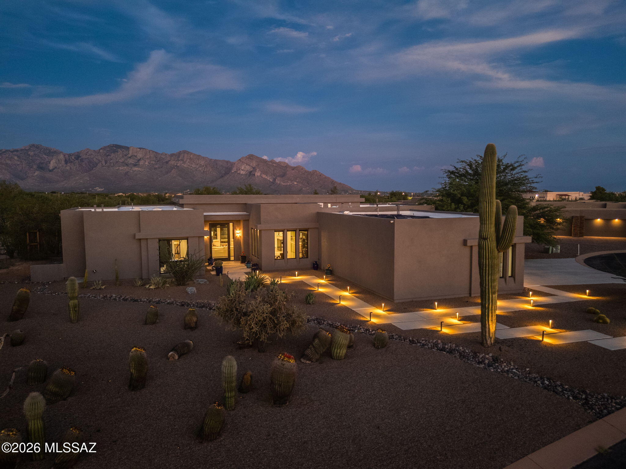 Outstanding Mountain Views! Experience refined desert luxury in the exclusive gated enclave of Sunset Canyon Estates in Oro Valley. Set on an expansive .84-acre homesite, this exceptional contemporary residence offers 4 BD/4BA 3355 sq ft of sophisticated smart-home technology, and designer finishes throughout. A dramatic illuminated walkway leads to an impressive 5ft by10ft pivoting glass entry door, creating a striking first impression. Inside, soaring recessed ceilings and walls of glass fill the expansive great room with natural light while framing captivating mountain and desert views. Designed for both elegant entertaining and everyday living, the spectacular gourmet kitchen features dual islands adorned with Alaska White granite and a Charcoal quartz waterfall island, complemented by top-of-the-line appliances and abundant custom cabinetry. The formal dining room showcases a stunning 450-bottle temperature-controlled wine vault, making every gathering memorable. The luxurious owner's retreat is a private sanctuary, complete with a spa-inspired bath featuring dual vanities, a soaking tub, an oversized walk-in tiled shower, and an expansive custom-designed closet. Two additional guest suites each offer private en-suite baths, while the fourth bedroom provides exceptional flexibility as an executive office or guest room. The resort-style backyard is designed to embrace Arizona's indoor-outdoor lifestyle. Relax on the elegant travertine patio surrounding the solar-heated pool and raised spa, accented by a decorative stone waterfall, custom fire pit, and built-in grilling station. Beyond the entertaining spaces, discover a tranquil botanical garden highlighted by a remarkable 250-year-old saguaro cactus and winding gravel pathways that invite peaceful walks through the beautifully preserved desert landscape. Luxury extends well beyond aesthetics with premium amenities including solid alder doors, automated window coverings, central vacuum, fire suppression system, whole-house surge protection, smart home control panel, advanced security features, and numerous additional upgrades. This extraordinary residence seamlessly blends timeless contemporary design, cutting-edge technology, and exceptional outdoor living with outstanding mountain views in one of Oro Valley's most desirable gated communities.