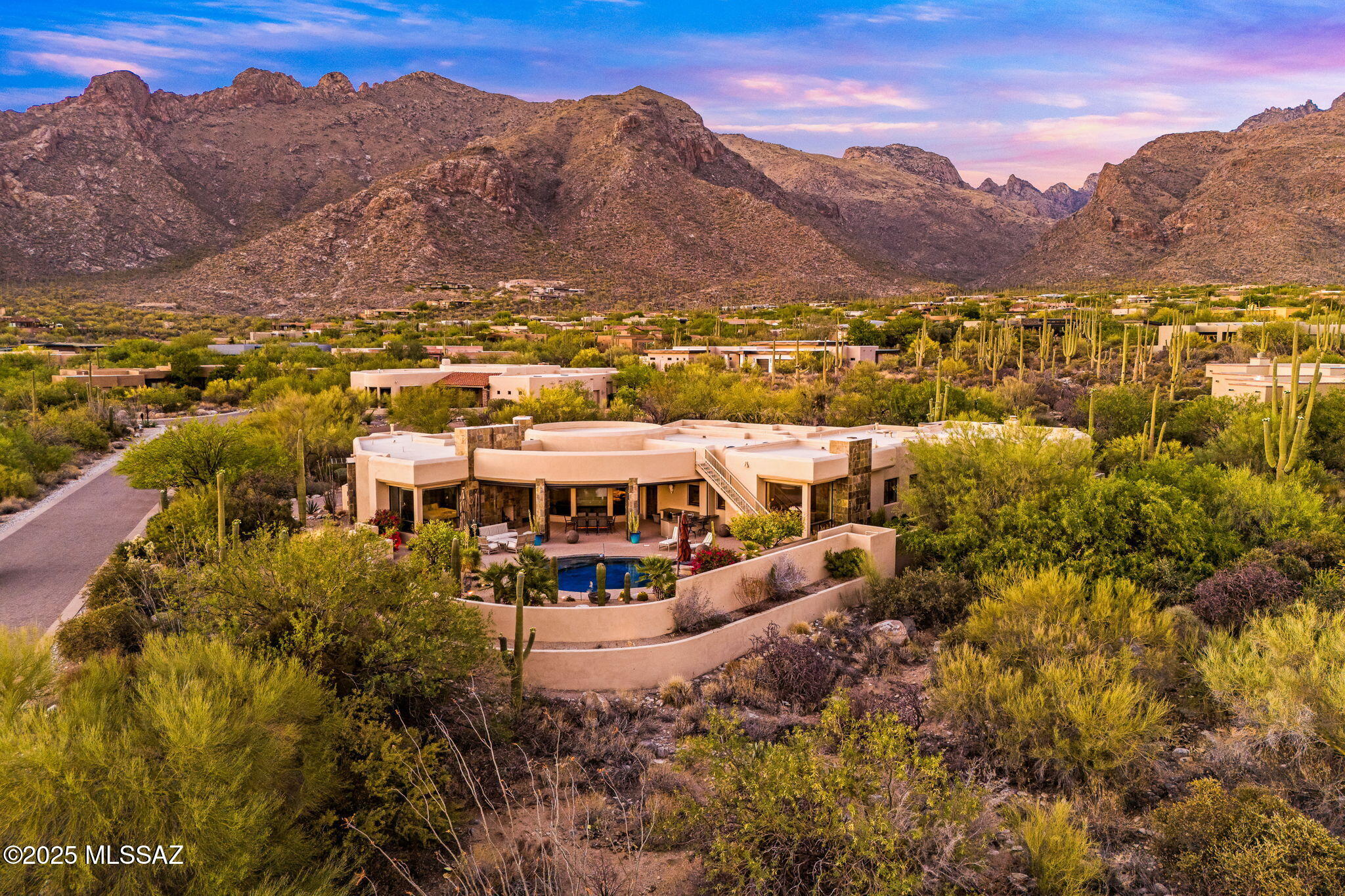 You will be awed by the striking design of this modern residence, where architecture and landscape exist in perfect harmony. Nestled within the prestigious double-gated sanctuary of Pima Canyon Estates, this 5,900 sq. ft. single-level home commands breathtaking city lights, dramatic Arizona sunsets, and majestic views of the Catalina Mountains. From the first approach, the custom glass entry door hints at what's inside: a space where clean lines, natural materials, and organic light define every room. The courtyard with a sculptural water feature sets the tone, leading guests through a tranquil transition from desert silence to architectural brilliance. Inside, an open-concept great room with soaring ceilings and a stone fireplace anchors the space. Large expanses of glass create walls that reveal panoramic mountain vistas. The living, dining, and kitchen areas flow effortlessly together, designed for large-scale entertaining.
The chef's kitchen offers top-of-the-line appliances, double ovens, a smooth electric cooktop, and a built-in wine refrigerator - designed for both culinary precision and social connection. An immense granite island invites guests to gather, with modern custom cabinetry with clean visual lines. Adjacent is a casual breakfast area overlooking the pool terrace and mountain views dotted with majestic saguaros.
This residence offers five ensuite bedrooms, each designed for comfort and privacy. The primary suite is a bespoke retreat, complete with a jetted soaking tub, dual LED-lit vanities, a walk-in rain shower opening to a glass-framed garden courtyard, and a custom walk-in closet. The second primary suite includes an impressive stone fireplace, a media room or lounge area, a custom walk-in closet, an in-suite beverage center, a rain shower, a jetted garden tub, and a private terrace entry near an outdoor fireplace ideal for guests or multi-generational living.
Step outside, and the home's connection to nature deepens. The resort-style backyard feels like an extension of the interior - a stage where the desert performs nightly. A custom pool and spa with architectural lighting create ambiance long after sunset. The built-in outdoor kitchen includes a BBQ, refrigerator, and ice maker, plus a rooftop view deck perfectly positioned for sunset dinners or entertainment. Multiple covered verandas and private seating alcoves provide space to unwind, entertain, or simply listen to the desert's quiet rhythm, viewing ancient saguaros, some centuries old.
Beyond its visual artistry, this estate delivers the ease of turnkey luxury. Offered fully furnished, every piece - from the designer sofas to the curated art that's been chosen to complement the architecture and desert palette. Smart-home technology, four fireplaces, an oversized three-car garage, and energy-efficient systems ensure both comfort and sustainability.

As day fades, the Catalinas blush pink, then violet, then indigo, and the home glows softly from within - where every evening feels like a private premiere, every moonrise a moment of awe.