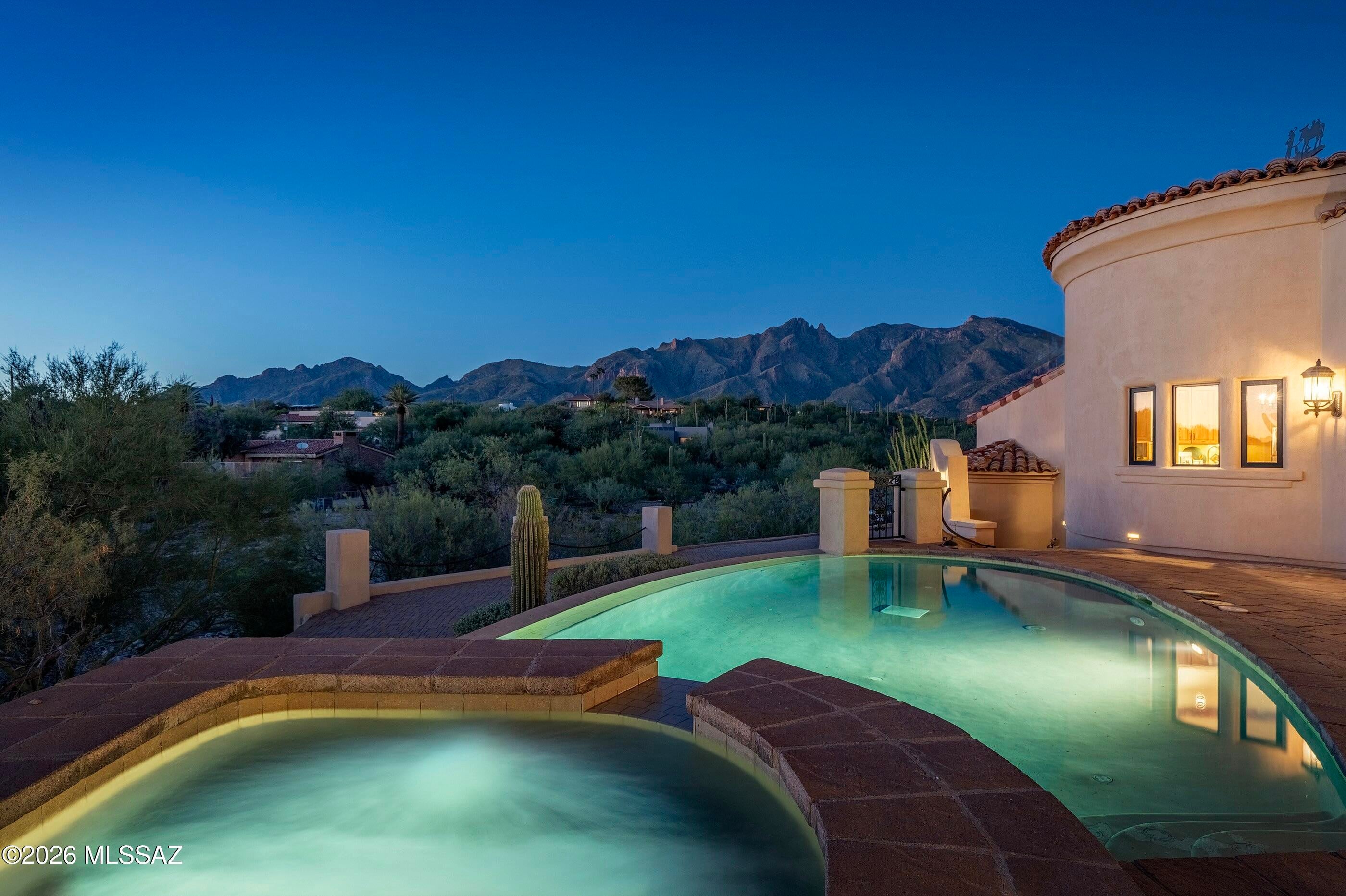 Set on more than an acre in the heart of the Catalina Foothills, this Spanish Colonial residence unfolds against a sweeping panorama of the Catalina Mountains. Soft stucco walls, clay tile rooflines, graceful arches, rounded forms, and iron detailing lend depth and character, while the pool and expansive outdoor living spaces connect the home to its remarkable desert setting. This 4,261 square foot residence offers four bedrooms, three and a half bathrooms, and a flex/media room. The residence lives primarily on one level, with subtle changes in elevation leading to the primary suite and garage. Handcrafted wood entry doors open to interiors defined by exposed timber beams, brick barrel vaults, arched passages, iron chandeliers, and a restrained palette of polished concrete and wood flooring.

The living room centers on a carved stone fireplace beneath warm wood beams. Glass doors open to an adjoining terrace, bringing the pool, surrounding vegetation, and mountain views into focus. Nearby, the formal dining room features a soaring ceiling, exposed beams, and a dramatic iron chandelier while remaining visually connected to the living room and kitchen.

Positioned between the living and dining rooms, the bar serves as both an architectural transition and a natural gathering place. Custom built in cabinetry, wine storage, refrigeration, granite surfaces, and counter seating create a distinct entertaining destination at the center of the home.

The kitchen pairs natural wood cabinetry with granite countertops, open display shelving, dual apron front sinks, stainless steel appliances, double wall ovens, and a professional style gas range set against a brick backdrop. A contrasting furniture-style island anchors the room, while a curved brick ceiling detail creates definition without separating it from the adjoining spaces.

The primary suite occupies a private wing with generous proportions and direct mountain views. Exposed beams, wood flooring, and a sitting area create a true retreat. The bathroom is enveloped in natural stone and features substantial dual vanities, a freestanding soaking tub, a walk-in shower with mosaic detailing, and an exceptionally generous fitted dressing room.

Three additional bedrooms are arranged separately, offering privacy for family and guests. Centrally positioned among them, the flex and media room includes custom cabinetry and a refreshment bar, with versatility as a screening room, secondary lounge, playroom, or informal gathering space.

Outside, columned loggias, covered dining areas, and expansive paver terraces extend the living spaces into the landscape. A built-in barbecue bar supports outdoor entertaining, while a fireplace defines an intimate seating area. The pool and spa follow the gentle curves of the residence, reflecting the sky and Catalina Mountains as mature saguaros frame the surrounding views.

Completing the property is an approximately 1,057 square foot three car garage, with a secondary bay featuring an oversized door for larger vehicles or recreational equipment. The central Foothills location offers convenient access to La Encantada, La Paloma Country Club, St. Philip's Plaza, the University of Arizona, Banner University Medical Center, Tucson Medical Center, and a broad range of dining, shopping, and everyday services. The result is a home that feels removed from the city when it should, and remarkably connected to it when it matters.