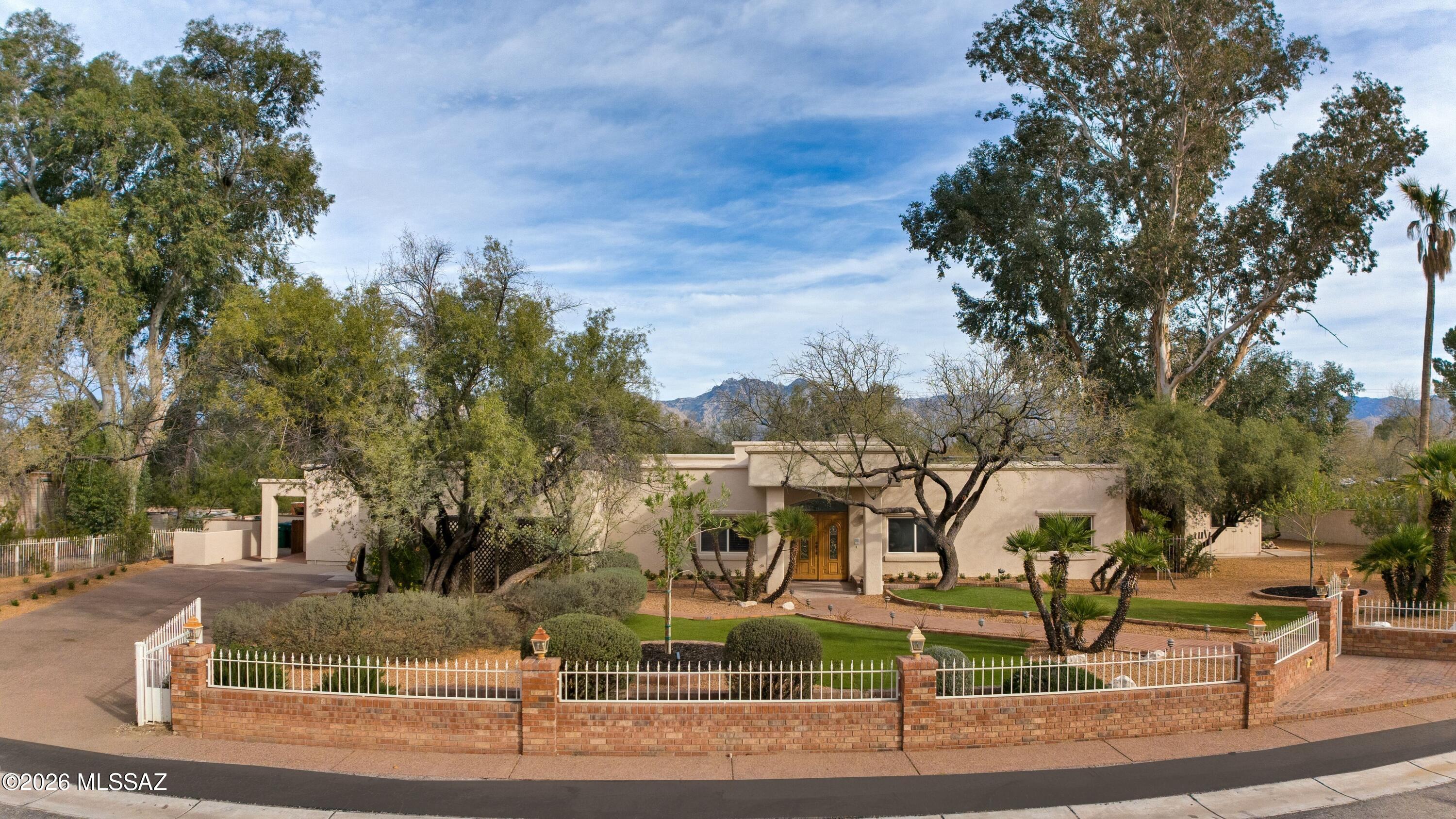 Introducing a rare opportunity within the prestigious, gated community of Tucson Country Club Estates. Offered for the very first time, this exceptional residence has been lovingly cared for by one family and is gracefully positioned on a lush and private .66-acre homesite framed by mature trees, gardens, and manicured low-maintenance turf, all secured by a charming brick with white iron fence accents and a gated driveway.
A welcoming covered porch with wood plank ceiling and double doors sets the stage for the warm, inviting interiors beyond. Inside, soaring ceilings, rich wood flooring, and timeless architectural details define the gracious living room, anchored by a gas fireplace and designed for entertaining and everyday comfort.
At the heart of the home, the expansive kitchen offers extensive cabinetry and dual granite islands; one functioning as a breakfast bar seamlessly opening to the cozy family room and sunlit breakfast nook, enhanced by a gas fireplace, creating an atmosphere of connection and warmth.

Thoughtfully designed for flexibility and privacy, the separate guest quarters include a living room, kitchenette, private bedroom, and bath, with direct access to both the backyard and driveway, ideal for guests, extended family, or multi-generational living. A spacious addition, previously enjoyed as a teen retreat, offers versatility for a game room, media lounge, or creative studio, complete with its own ½ bath.

The private bedroom wing features a handsome den with custom built-in bookcases and a desk, three generously sized bedrooms, and a serene primary suite with its own fireplace, walk-in closet, and spacious bath featuring dual split vanities, a soaking tub, and a separate shower.

The backyard is a true sanctuary, highlighted by a sparkling pool, a rock waterfall, an attractive three-tier fountain, and multiple fenced areas for recreation. A north-facing covered patio and a new built-in stainless-steel barbecue create the perfect setting for outdoor gatherings year-round.

Additional amenities include a detached, climate-controlled fitness room, abundant storage, and expansive side yards.
Tucson Country Club Estates is celebrated for its welcoming sense of community, offering neighborhood gatherings, parades, and peaceful streets ideal for walking and enjoying the surrounding beauty. With no HOA, this is a rare offering that combines privacy and an exceptional lifestyle.